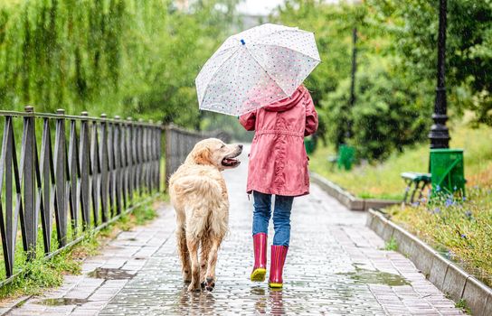 Rear view of little girl walking dog under rain along wet alley