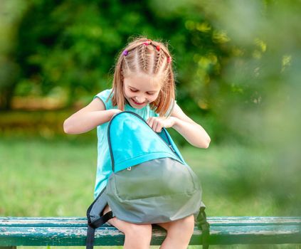 Schoolgirl peeking into open backpack on bench in park