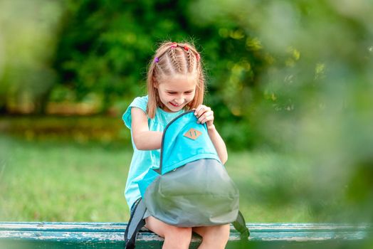 Schoolgirl peeking into open backpack on bench in park