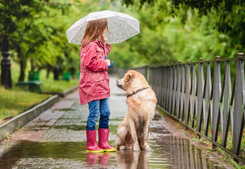 Little girl and golden retriever dog walking along park alley under rain