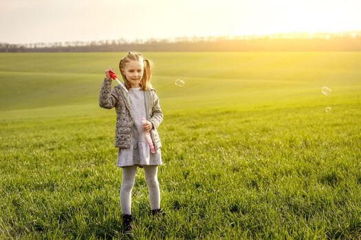 Cute little girl blowing soap bubbles on green field in spring