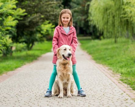 Little girl standing with golden retriever dog between feet on paved alley in park