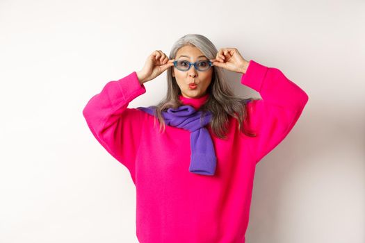 Trendy asian grandmother in sunglasses, pucker lips and looking surprised at camera, staring amazed at special offer, standing over white background.