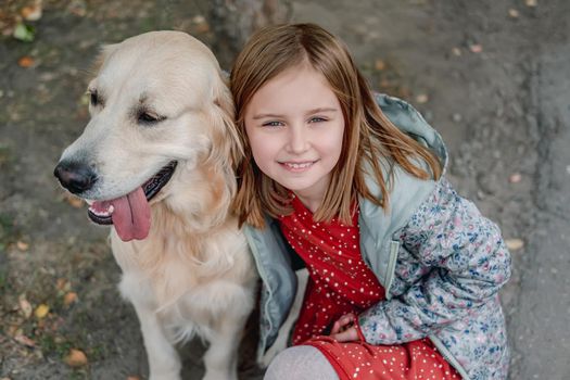 Little girl hugging golden retriever dog while looking up at camera on autumn street, top view