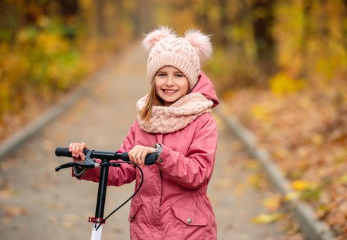 Little girl with a scooter standing on road in autumn park and smiling at camera