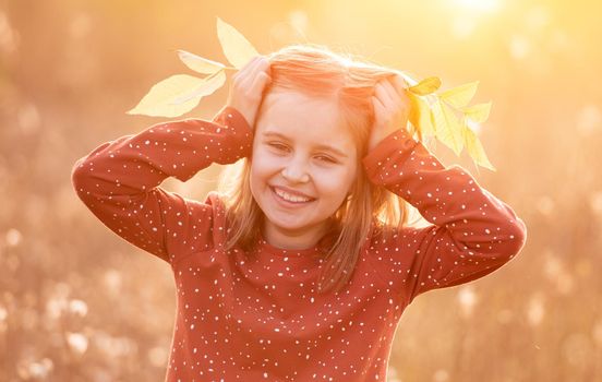 Portrait of smiling little girl holding autumn leaves on head in sunlight