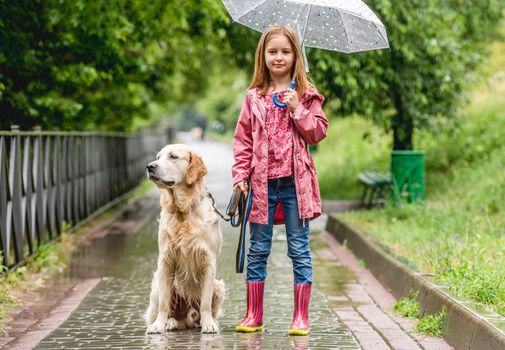Little girl with umbrella walking golden retriever dog in rainy park