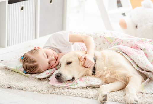 Happy little girl sleeping with lovely dog under blanket on floor in light room