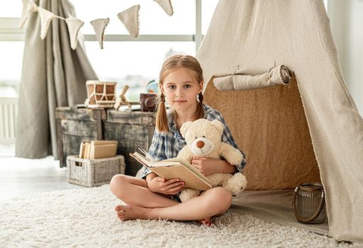 Nice little girl with bear and paper book sitting on room's floor decorated with wigwam, chest and retro lantern