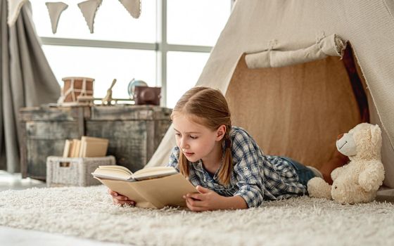 Cute little girl reading adventure book lying in wigwam settled in kids room, panoramic orientation