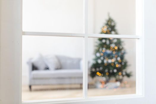 Blurred Christmas tree decorated with shining garland placed near gray couch in room through window