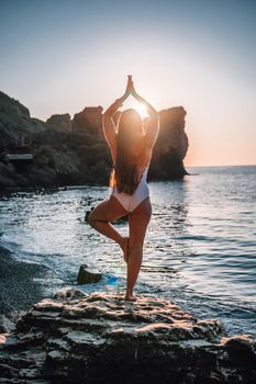 Young woman in swimsuit with long hair practicing stretching outdoors on yoga mat by the sea on a sunny day. Women's yoga fitness pilates routine. Healthy lifestyle, harmony and meditation concept.