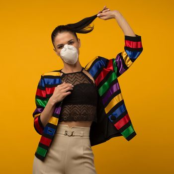 Beautiful slim girl fashion model posing in a protective respirator on a yellow background. LGBT community rainbow color jacket