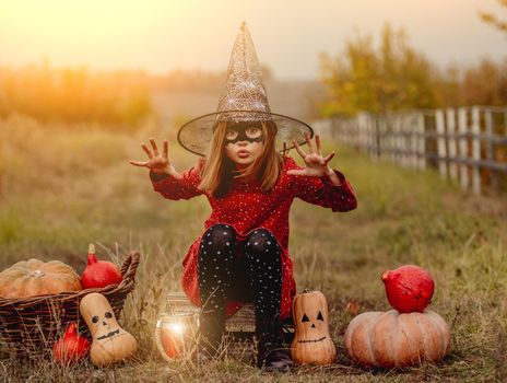 Little girl in halloween costume sitting next to pumpkins while frightening at camera on autumn nature