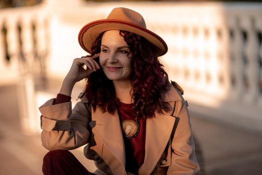 Outdoor fashion portrait of young elegant fashionable brunette woman, model in stylish hat, choker and light raincoat posing at sunset in European city