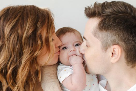 Young man and woman kissing lovely baby in cheeks while standing on white background