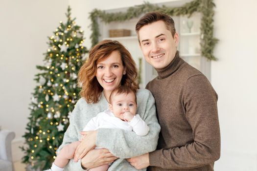 Cheerful man and woman holding baby and looking at camera while standing on background of stylish room and Christmas tree