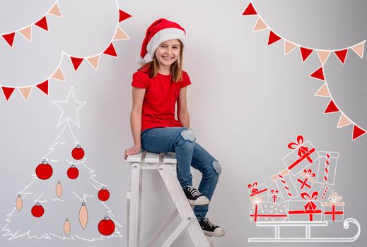 Happy little girl in santa hat sitting on ladder in middle of christmas drawings on white wall background