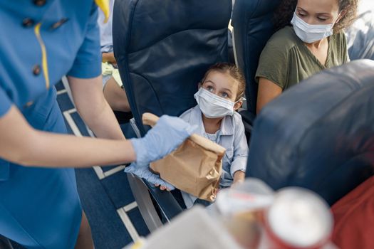 Cute girl in protective face mask looking at flight attendant serving lunch to little passenger on board. Traveling by airplane during Covid19 pandemic. Selective focus