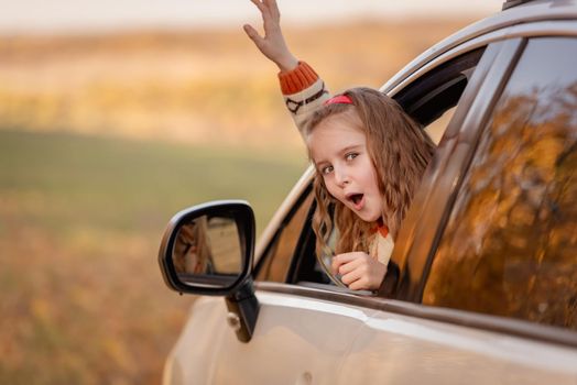 Happy little girl raising hand in car window on autumn background