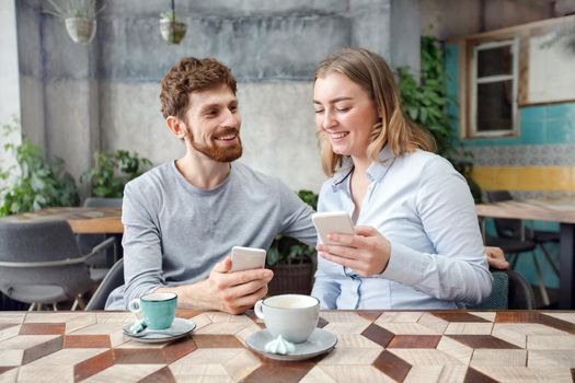 Young smiling man and woman with phones having coffee at table in modern coffee shop
