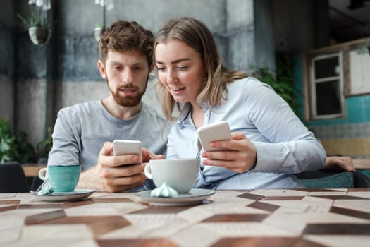 Young couple interacting while using smartphones and drinking coffee in shop.