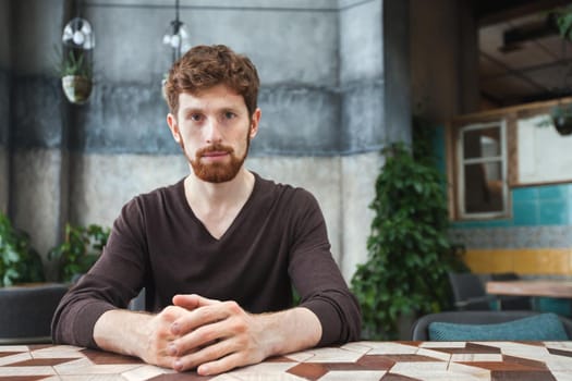 Portrait of young bearded man sitting confidently at table and looking at camera in a public place