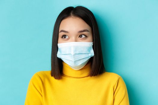 Covid-19, social distancing and pandemic concept. Headshot of cute asian woman with short dark hair and medical mask, looking left, standing over blue background.