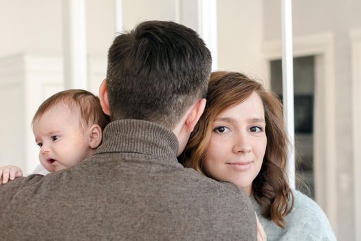 Back view of young man embracing beautiful woman and sweet baby while standing in cozy room together
