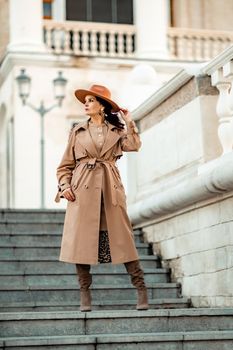 Outdoor fashion portrait of young elegant fashionable brunette woman, model in stylish hat, choker and light raincoat posing at sunset in European city