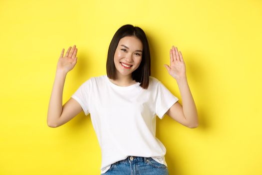 Friendly young asian woman saying hello, raising empty hands up and smiling, greeting you, standing over yellow background.