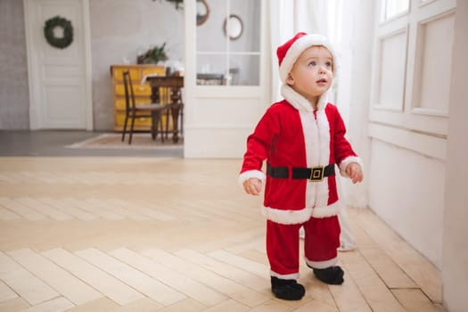 Cute little boy in red and white costume of Santa Claus standing on parquet and looking aside on background of dining room