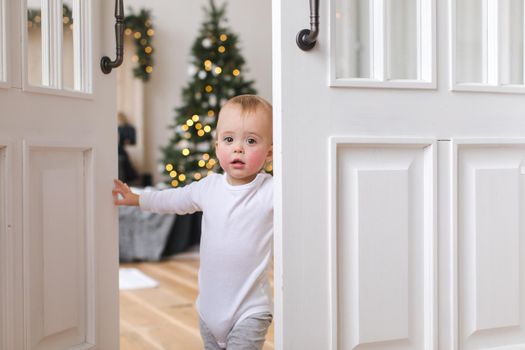 Adorable infant standing in doors looking at camera on background of Christmas tree in bedroom.