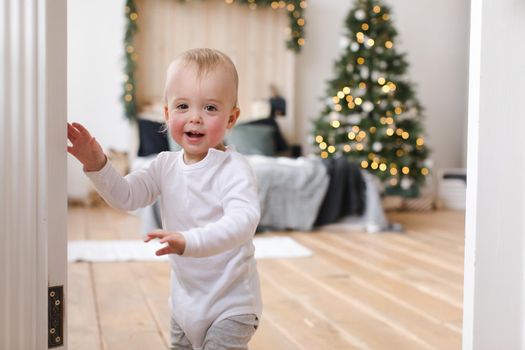 Adorable infant standing in doors looking at camera on background of Christmas tree in bedroom.