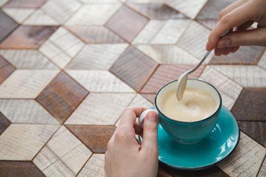 Cup of cappuccino with milk foam on wooden table. Female hand holds spoon with sugar