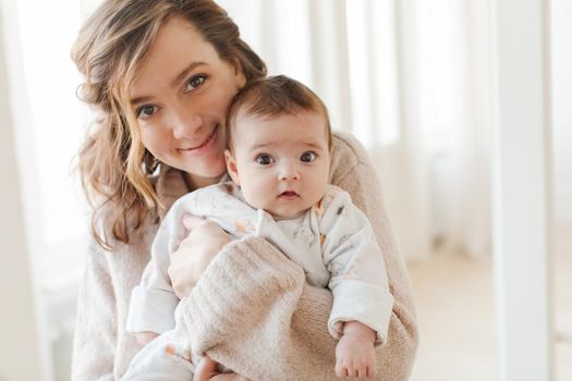 Beautiful young woman smiling and looking at camera while standing in light room and holding charming baby