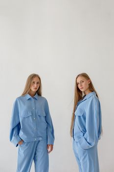 two young twin sisters with long blond hair posing on white background in oversize clothes with bare feet and looking at camera. They wear blue suits that look like pajamas. Stylish fashion photoshoot