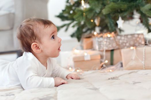 Side view of charming infant baby crawling on floor near Christmas tree with presents looking curious