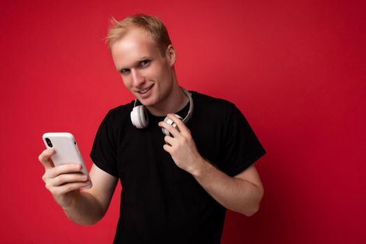 Photo of handsome good looking blonde young man wearing black t-shirt and white headphones standing isolated on red background with copy space holding smartphone and surfing internet on mobile phone looking at camera.