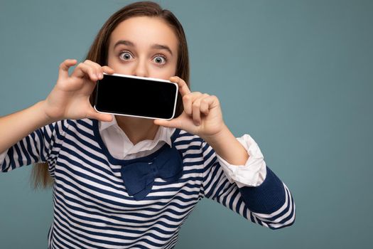 Closeup portrait of beautiful surprised brunette girl wearing striped longsleeve standing isolated on blue background with copy space holding smartphone showing phone in hand with empty screen display for mockup pointing at gadjet looking at camera.