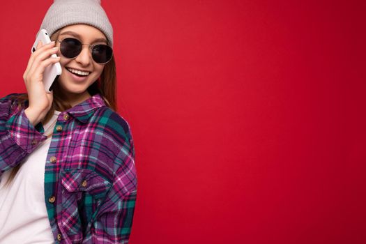 Closeup photo of beautiful happy positive young blonde woman wearing hipster purple shirt and casual white t-shirt grey hat and sunglasses isolated over red background holding in hand and talking on mobile phone looking to the side. Copy space