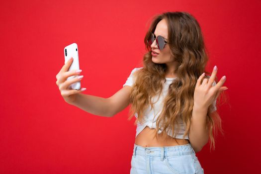 young woman holding mobile phone taking selfie photo using smartphone camera wearing everyday stylish outfit isolated over colorful wall background looking at device screen