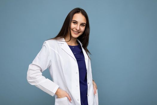 Photo of young attractive beautiful positive smiling brunette woman with sincere emotions wearing white medical coat isolated over blue background with empty space.