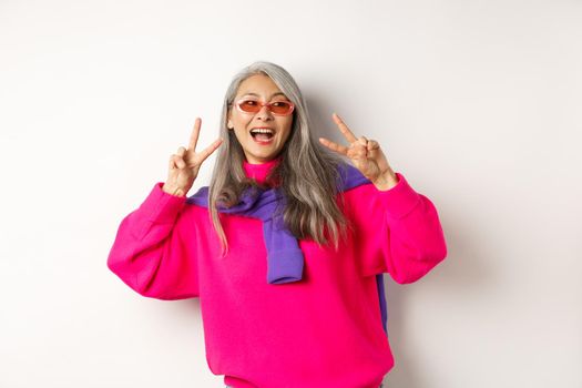 Fashion and beauty concept. Image of stylish asian senior woman in sunglasses smiling, showing peace signs and looking happy, standing over white background.