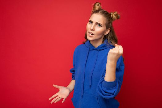 Photo of emotional agressive young beautiful blonde woman with two horns with sincere emotions wearing hipster bright blue hoodie isolated over red background with free space and showing fist.