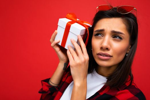 Closeup photo of beautiful emotional young brunet woman isolated on red background wall wearing white casual t-shirt and stylish red and black shirt holding white gift box with red ribbon.