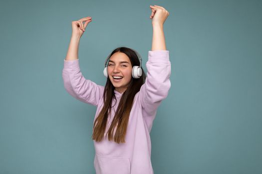 Attractive positive smiling young brunette woman wearing light purple hoodie isolated over blue background wall wearing white bluetooth headphones listening to good music and dancing looking at camera.