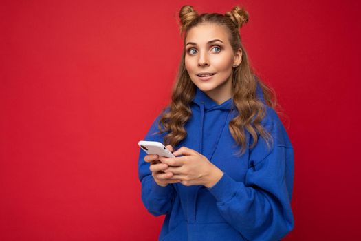 Attractive young blonde woman wearing blue stylish hoodie standing isolated over red background texting message via phone looking to the side.