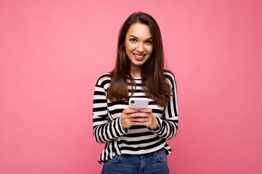 Beautiful young happy brunette woman using mobile phone communicating via texting message wearing sweater isolated on wall background looking at camera.