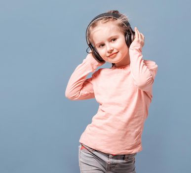 Cute little girl with headphones listening to music on light blue background. Copyspace